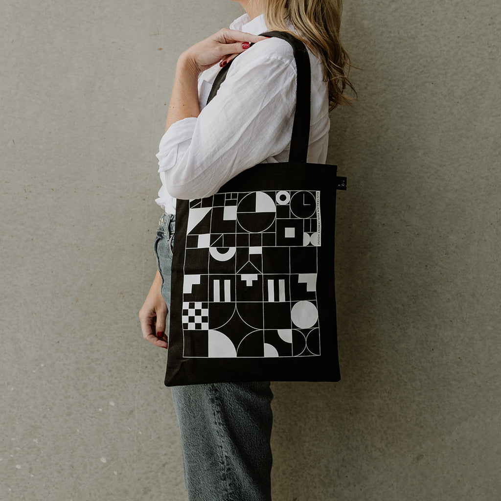 Side view of a woman in a white shirt wearing a black Parliament Shop branded tote bag with a architectural glyph pattern