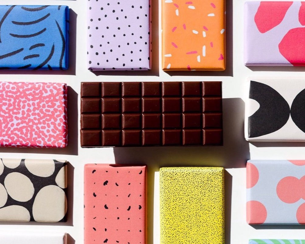 Flat lay of ten colourful wrapped chocolate blocks in multiple colours and one unwrapped chocolate block in the middle.
