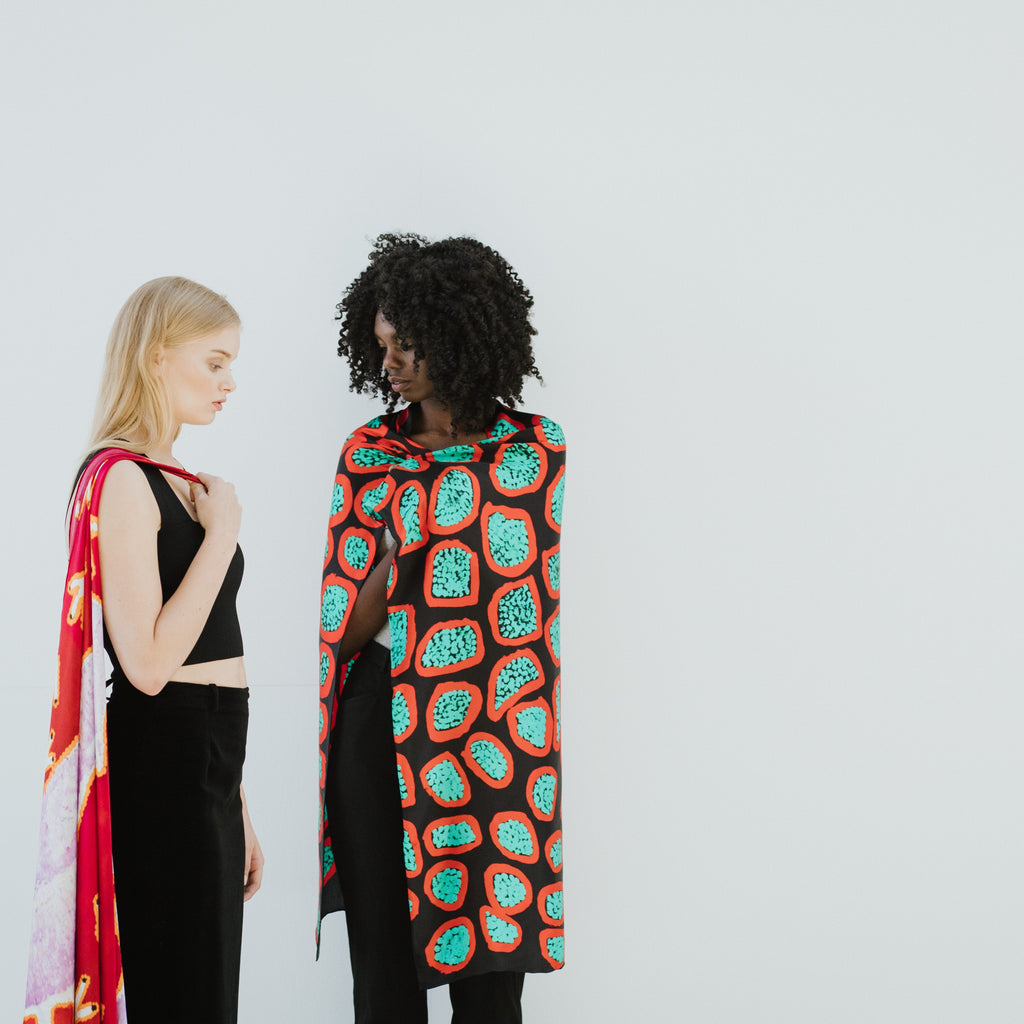 Two women standing against a white background with One of Tweleve coloured patterned scarves draped over their shoulders.