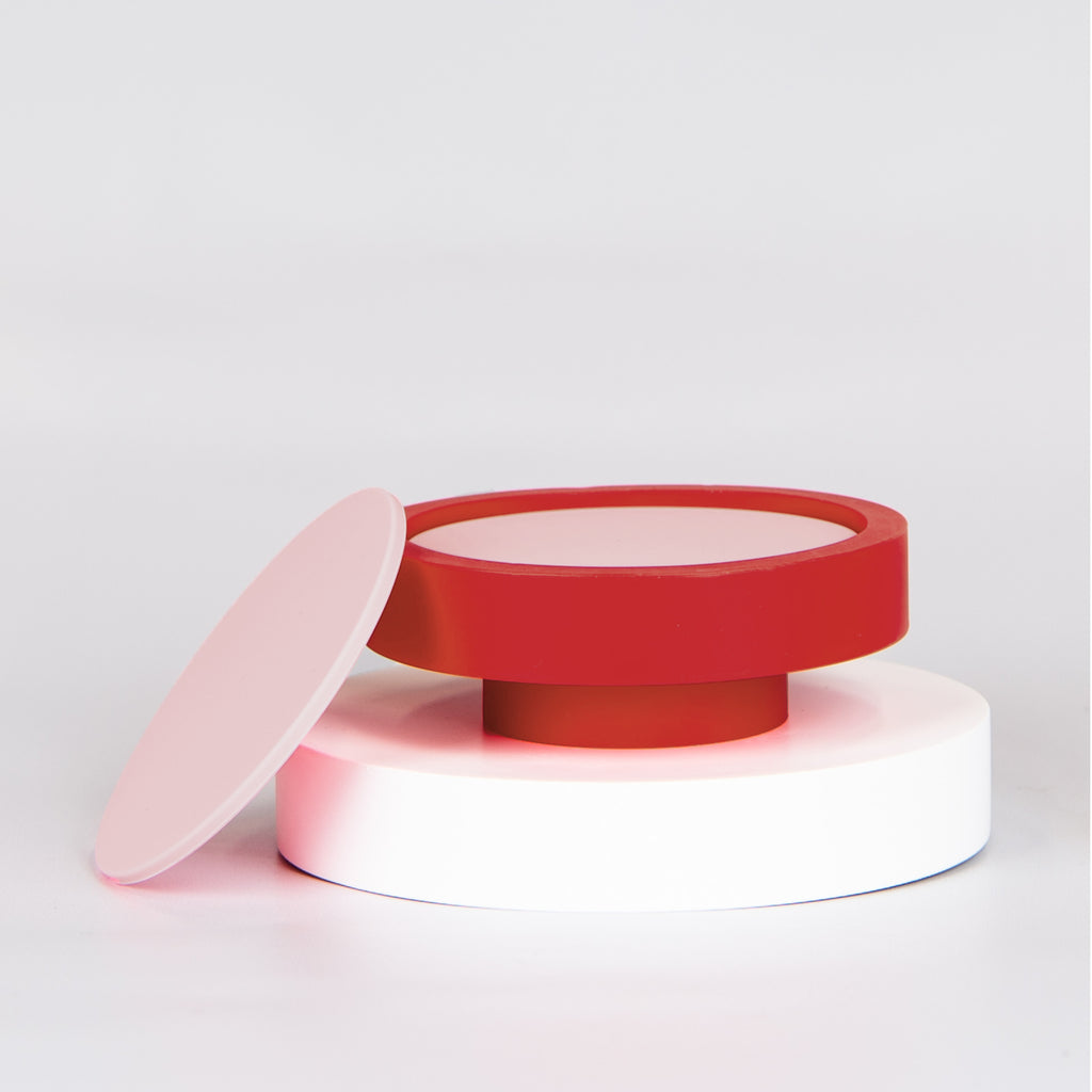 Round pink silicone coasters in a red base on top of a small round circle plinth with one coaster leaning against the side.