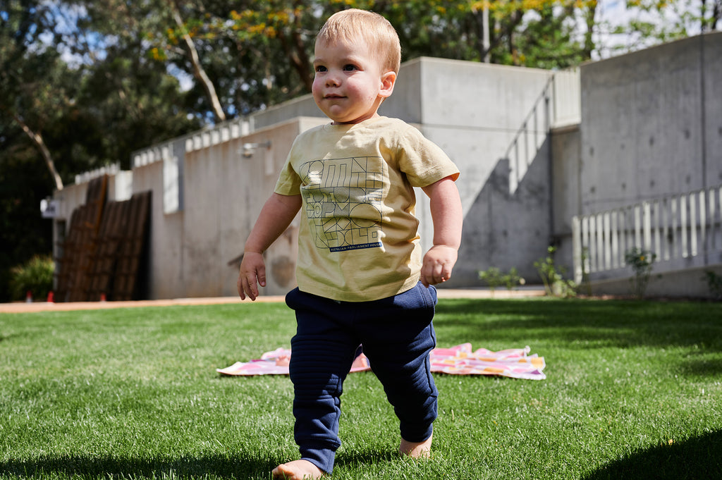 toddler walking on grass wearing mustard yellow t-shirt and navy pants