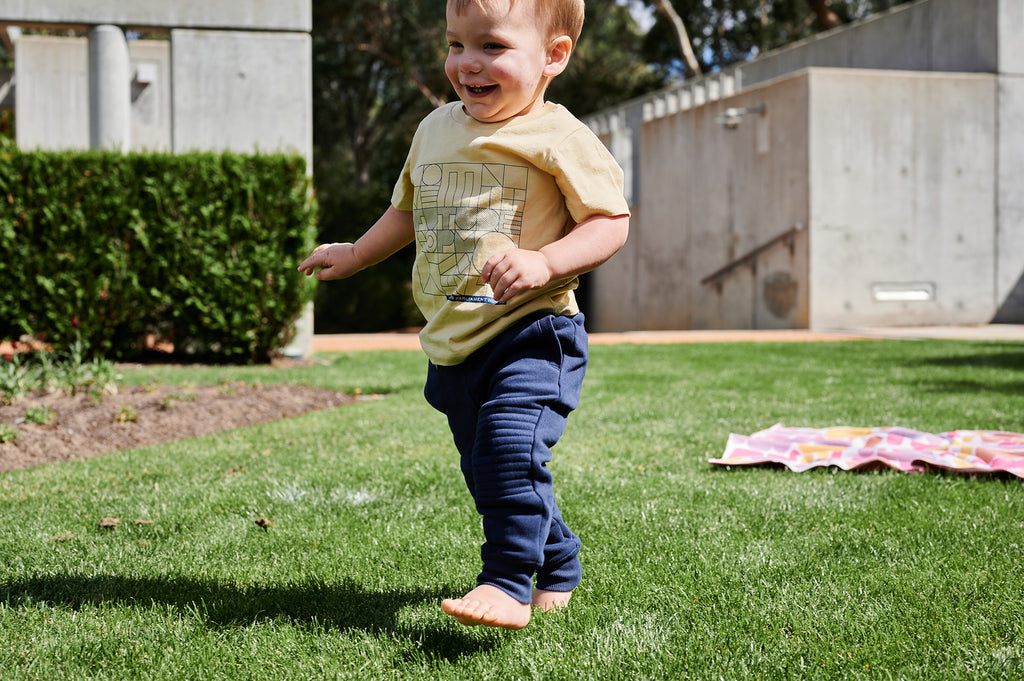 toddler smiling walking on grass wearing mustard yellow t-shirt and navy pants