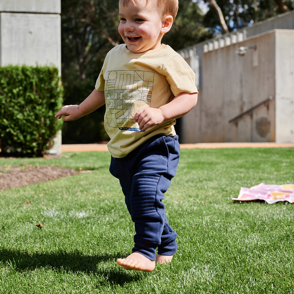 toddler smiling walking on grass wearing mustard yellow t-shirt and navy pants