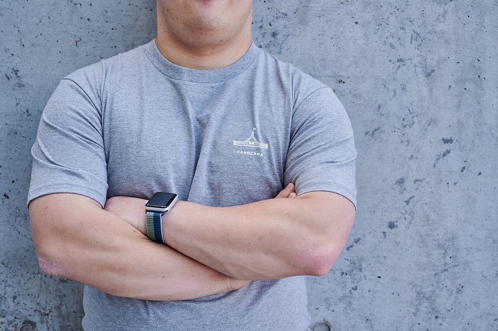 Man with arms crossed wearing a light grey t-shirt with the Parliament House logo on it and the words 'Canberra'.