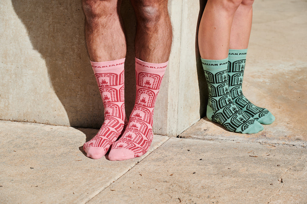 two pairs of feet standing against different sides of a wall. One is wearing pink socks and the other green socks