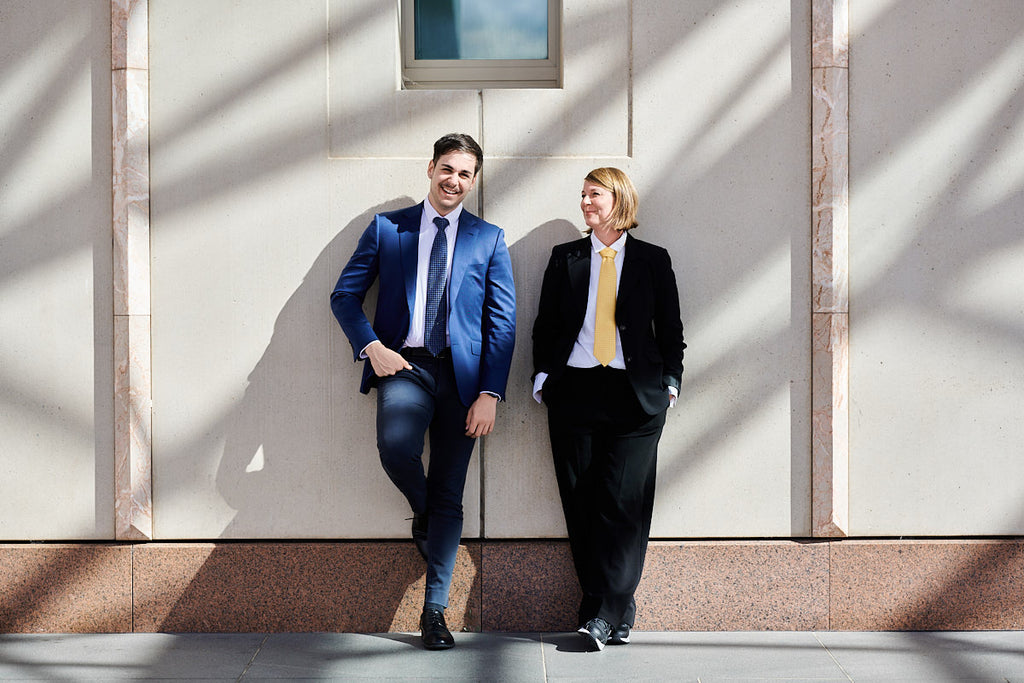 Man and woman wearing suits and a tie standing against a wall