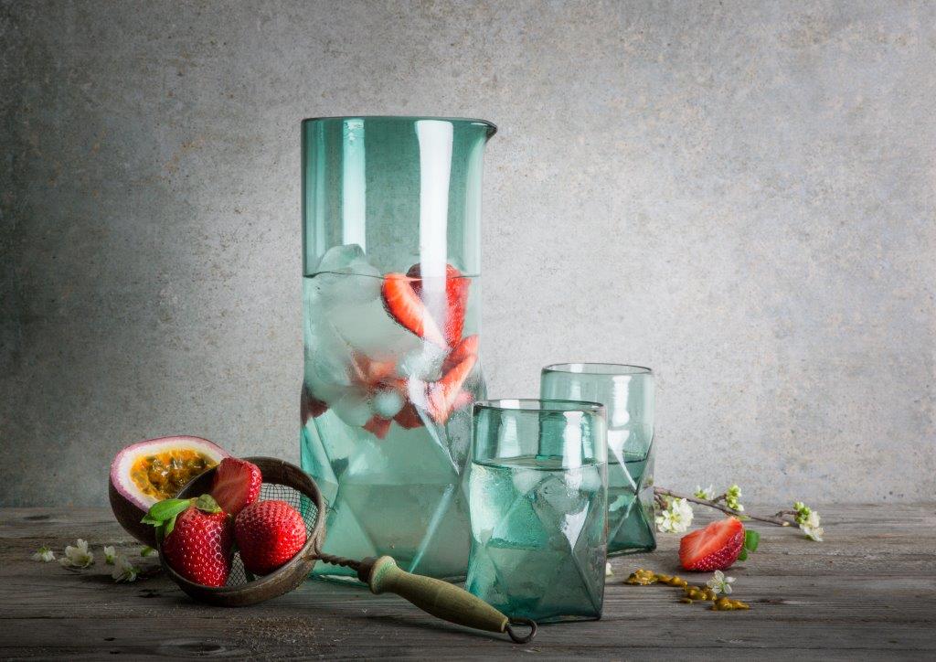 Blue Clear Glass Jug with Iced Water and Sliced Strawberries, surrounded by matching cups, passionfruit and strawberries