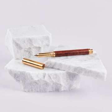A ballpoint pen with a wood barrel and brass cap and trims arranged on pieces of marble.