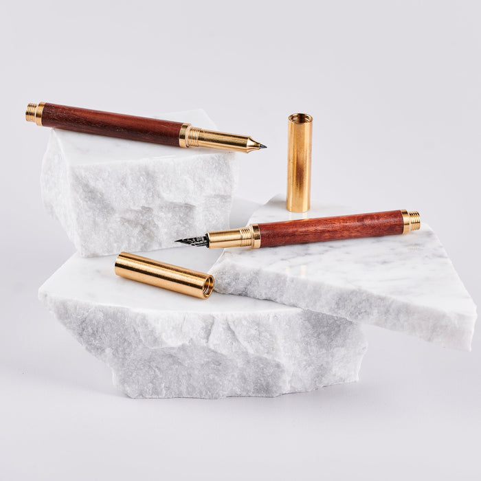 Two pens with wood barrels and brass caps and trims arranged on pieces of marble. 