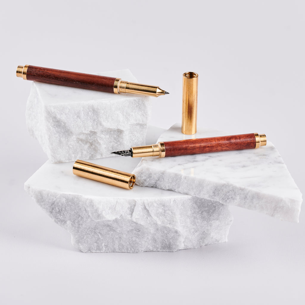 Two pens with wood barrels and brass caps and trims arranged on pieces of marble. 