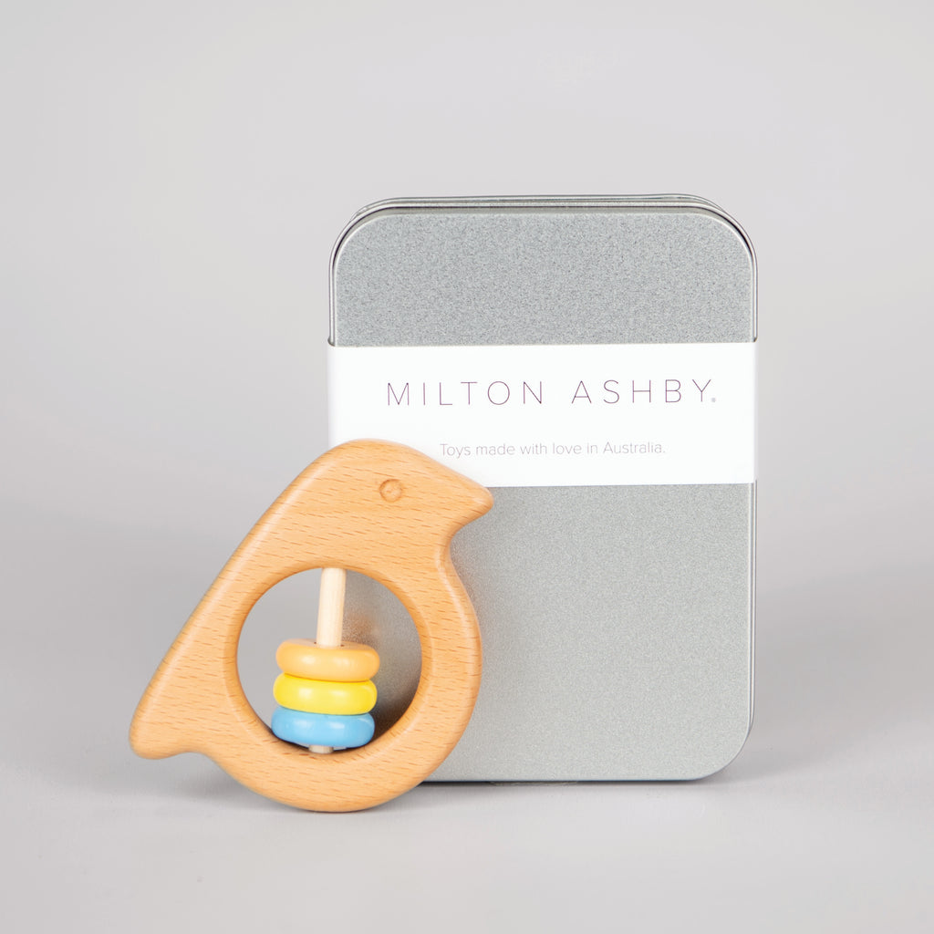 Wooden bird rattle toy in front of metal box with the words 'Milton Ashby'.