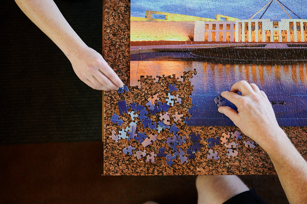 two hands completing the bottom left corner of a jigsaw puzzle of parliament house on a table
